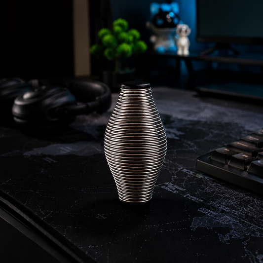 A ribbed metallic silver vase standing on a dark desk in a dimly lit room with a keyboard, headphones, and a monitor in the background.