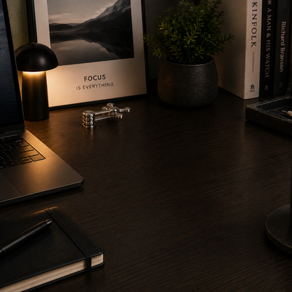 A dark wood desk styled with a laptop, notebook, pen, small lamp, framed print, books, a candle, and a potted plant in a dimly lit home office setting.